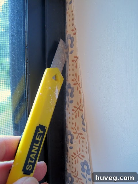 Hand using a utility knife to carefully cut wallpaper along a window frame.