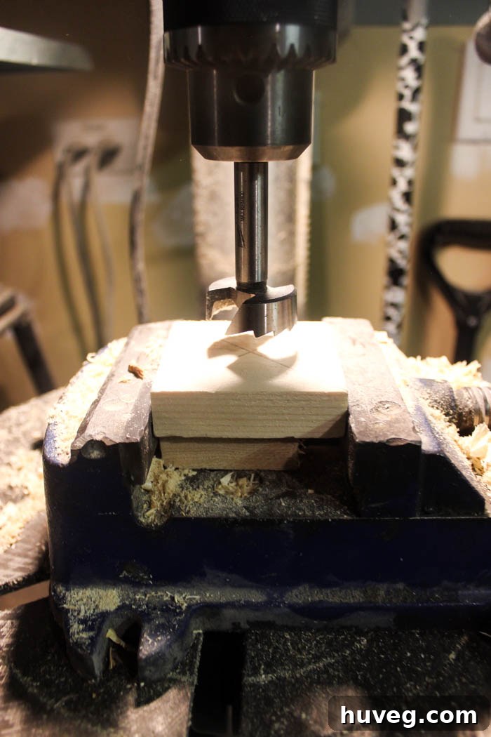 A drill press in action, with a Forstner bit drilling through a clamped wood block and a sacrificial board underneath.