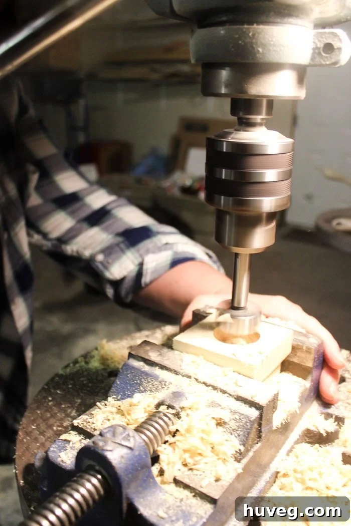 A drill press shown from the side, demonstrating the alignment of the Forstner bit with the center point on the wood block before drilling.