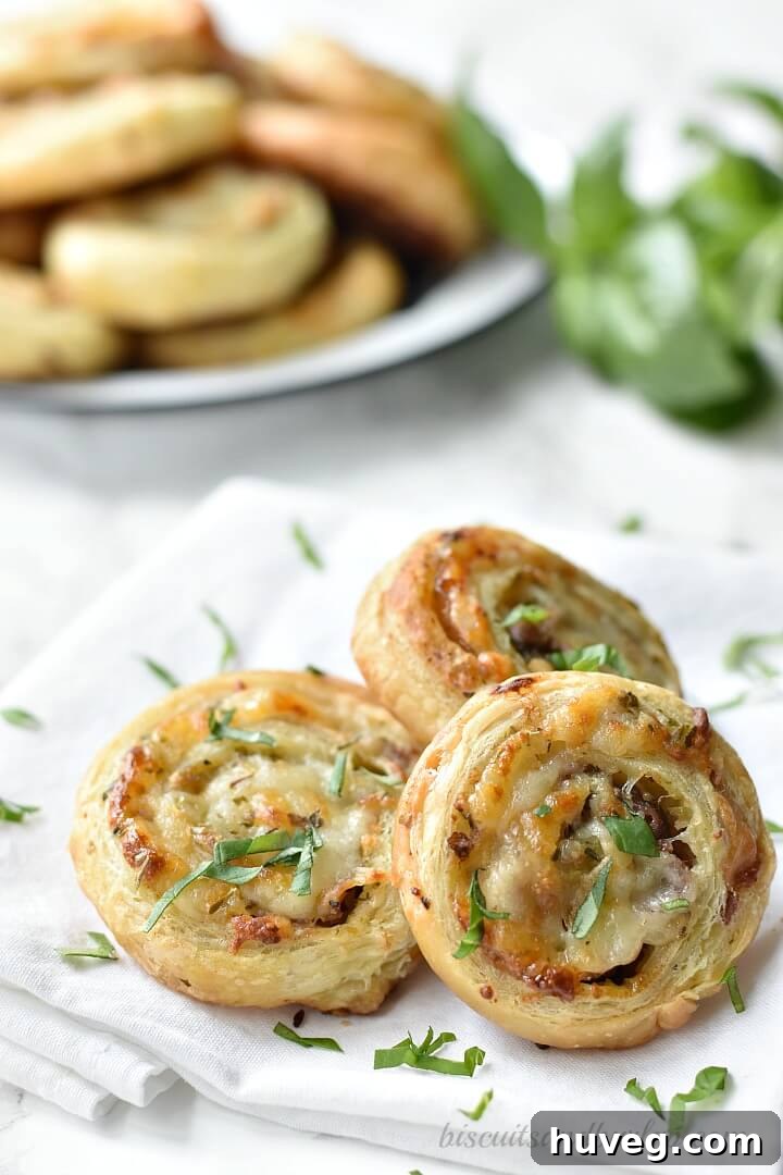 Close-up of golden brown puff pastry pinwheels on a white napkin, with a plate of various appetizers blurred in the background, hinting at a lively party setting.
