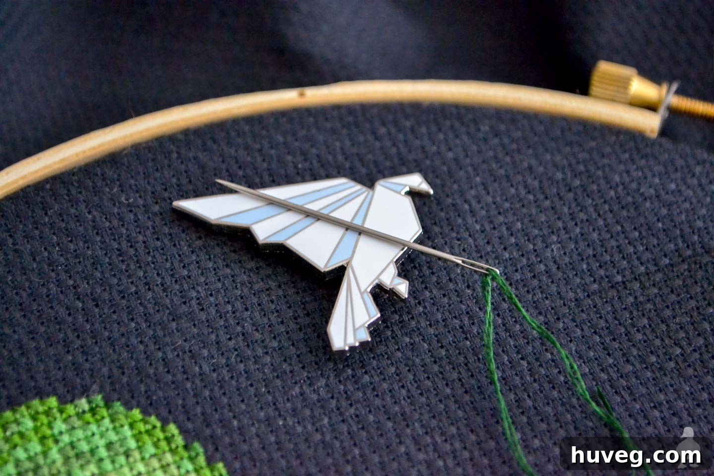 A magnetic needle minder, featuring a white origami bird, positioned on black Aida cross stitch fabric.