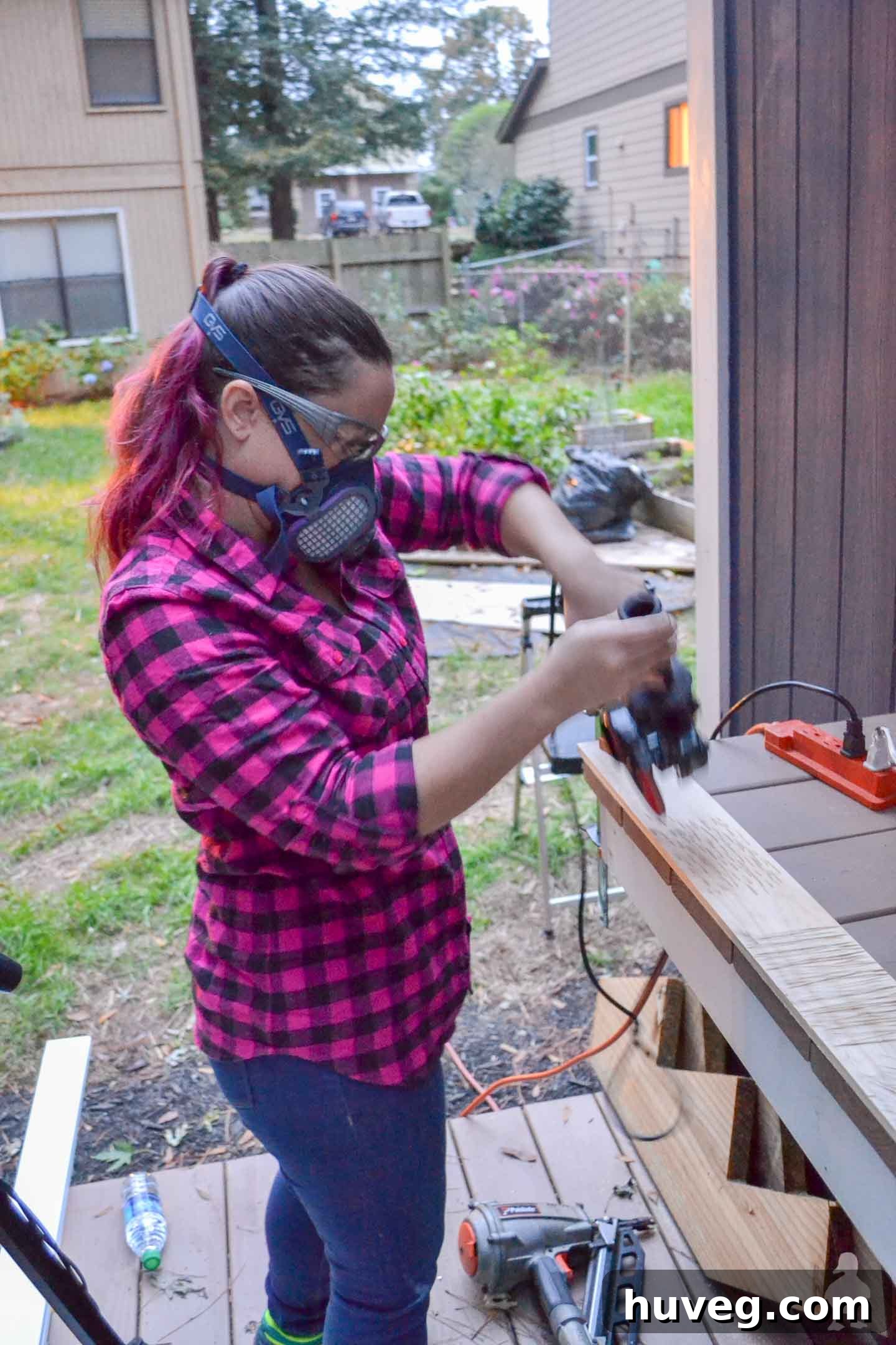 A woman in a pink plaid shirt uses an angle grinder to carve intricate textures into a piece of scrap wood, demonstrating the power carving technique for DIY rustic ornaments.