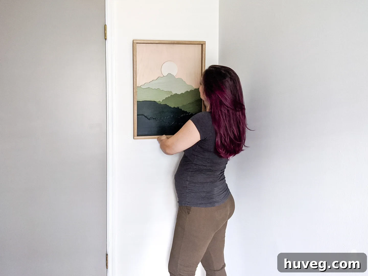 A woman with a bun on her head, possibly Sarah Fogle, carefully hangs a piece of scroll saw art featuring green mountains on a pristine white wall in a nursery. The art is framed in natural wood, adding a touch of rustic charm to the room.
