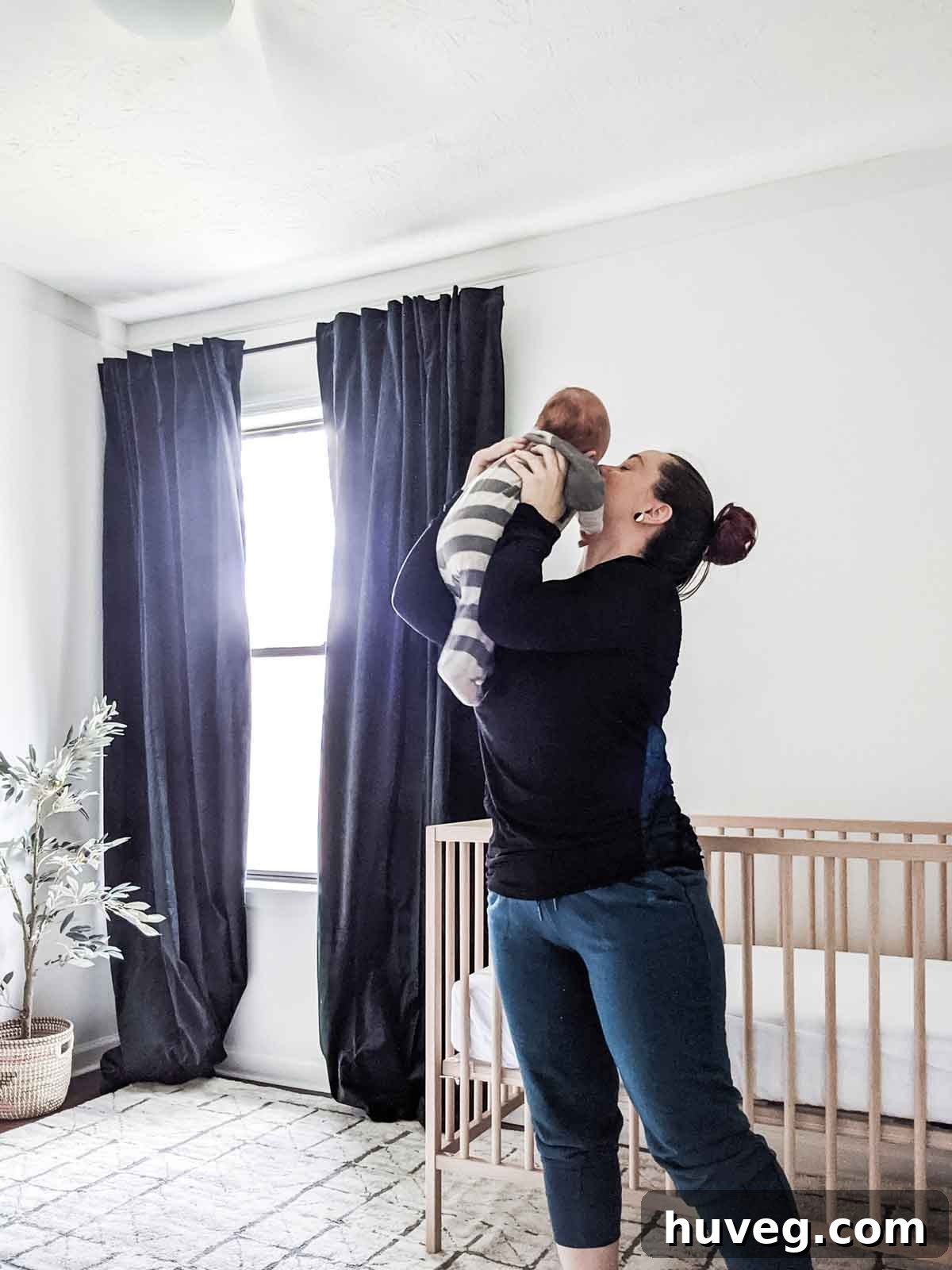 A woman with her hair pulled back in a bun cradles her baby inside a serene nursery. The room features clean white walls and elegant dark green curtains, creating a calming atmosphere for the child.