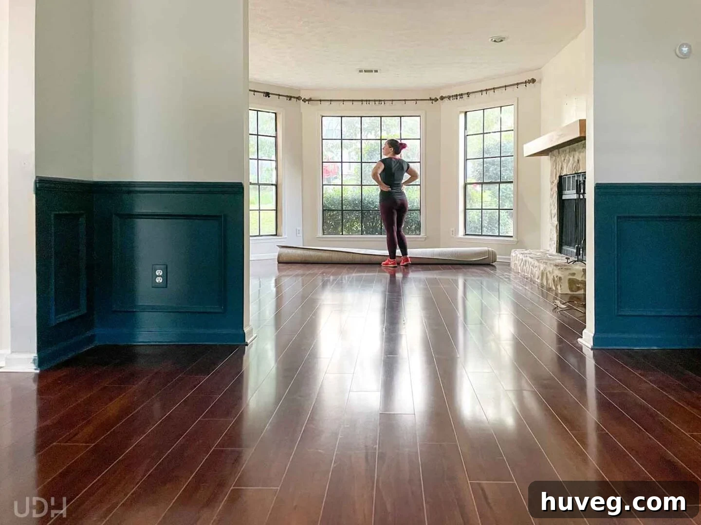 Sarah Fogle stands thoughtfully in front of her emptied living room windows. The curtains have been removed, and the rug is rolled up, symbolizing a significant transition as she prepares to sell her house and embark on a new adventure.