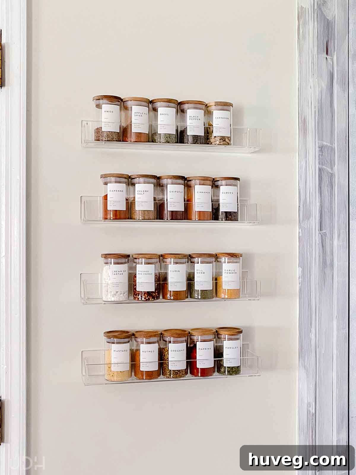 A clear acrylic spice rack filled with elegant glass spice jars, featuring wooden tops and white labels, beautifully organized in the kitchen.