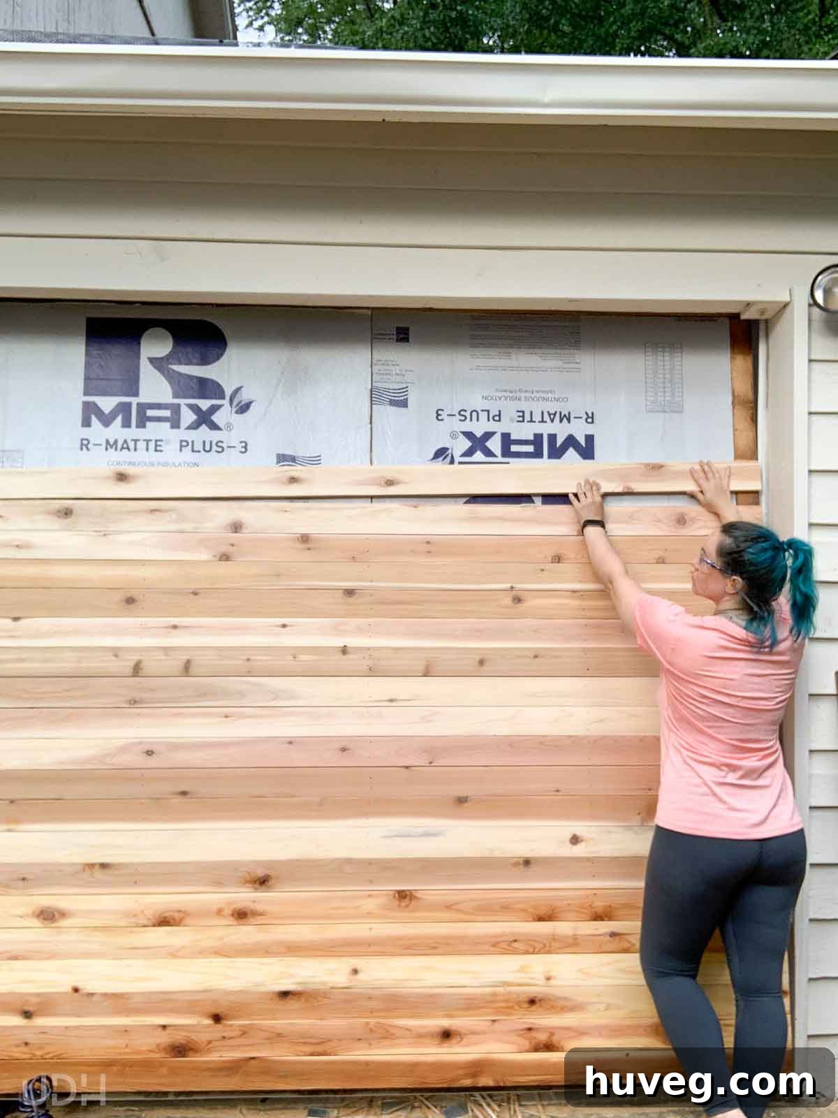 A woman in the process of installing handmade tongue and groove boards onto a garage door, with visible insulation, during its transformation.