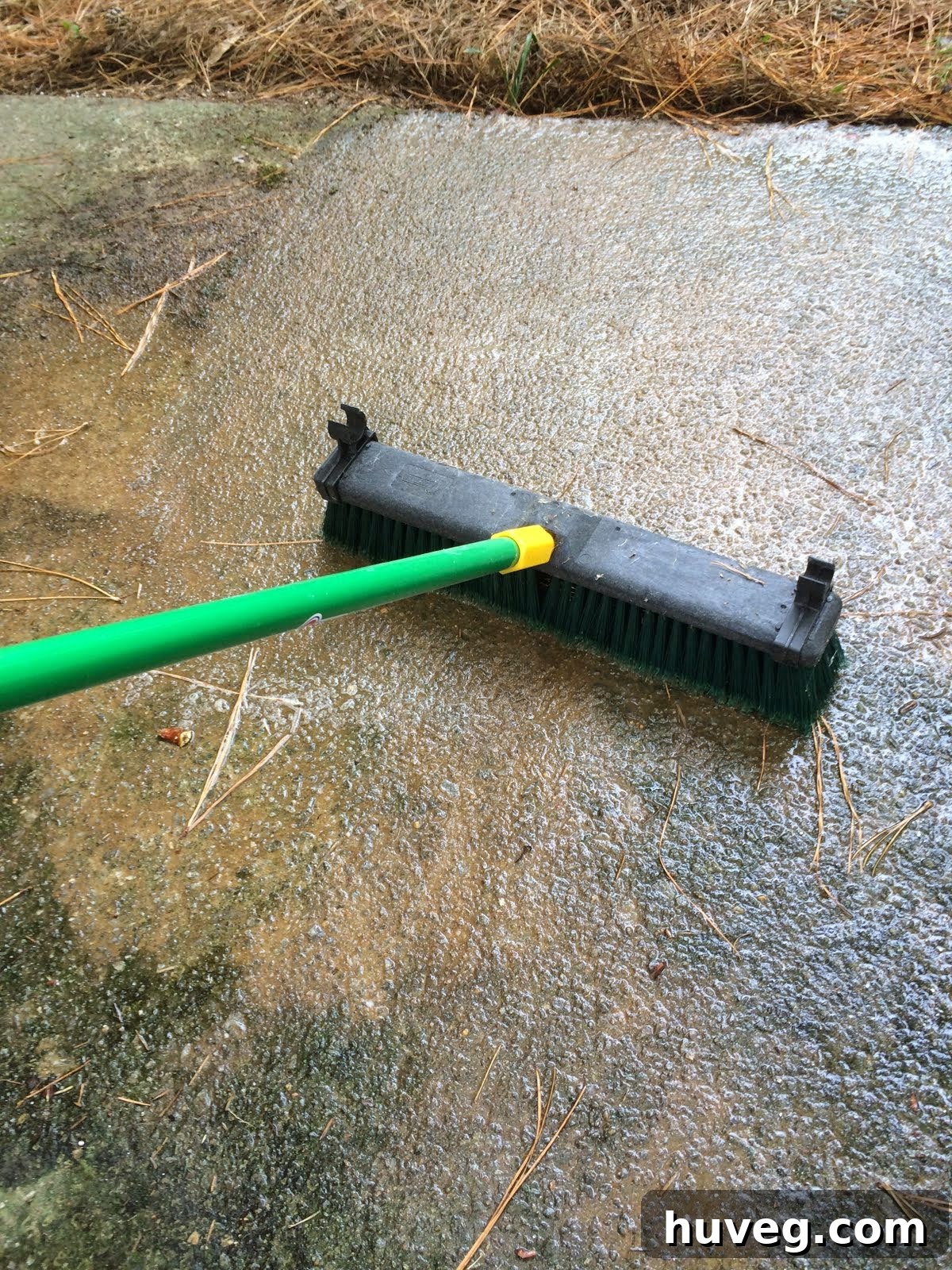 Using a long-handled stiff brush to scrub deck cleaner into a dirty patio surface