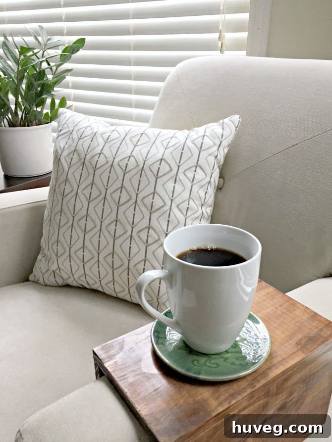 Coffee resting on a DIY sofa armrest tray table next to a white patterned pillow and houseplant, in a cozy living room.