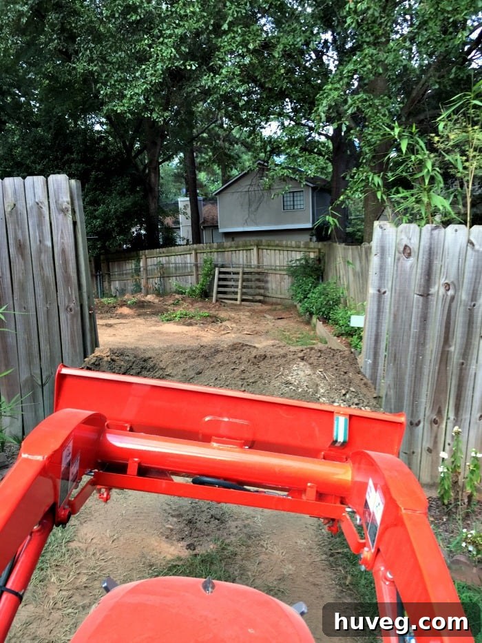 Operating a backhoe with a full load of dirt, carefully fitting through a narrow fence opening.